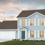 A two-story light blue house with white shutters, a dark front door, and a double garage sits on a manicured lawn with small shrubs and flowers—a stunning example of custom homes Gainesville at sunset under a partly cloudy sky.
