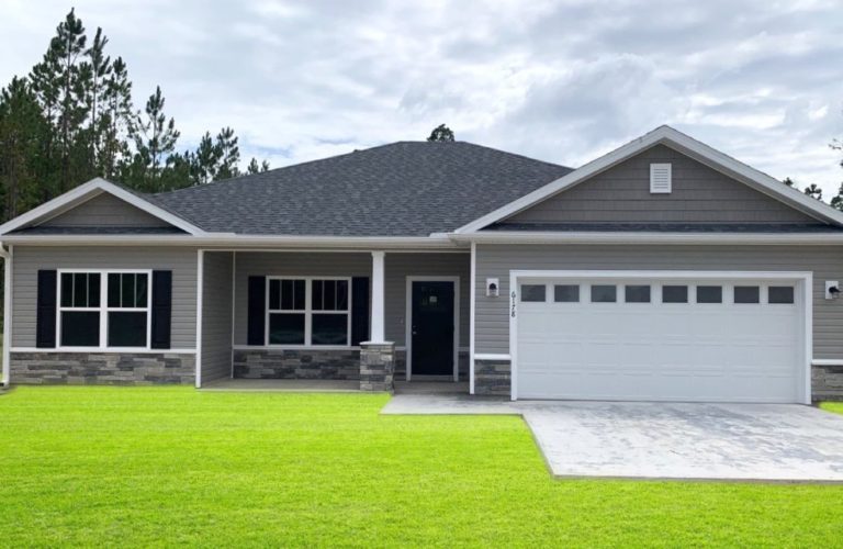 A single-story suburban house with gray siding, black shutters, and a two-car garage—perfect for those seeking custom homes in North Central Florida. The front yard boasts neatly cut green grass, a concrete driveway, and tall trees under a cloudy sky.