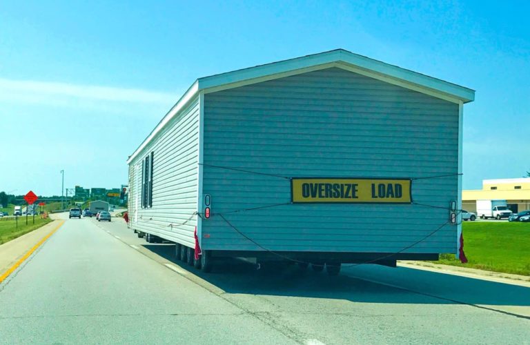 A large custom home is being transported on a highway in North Central Florida, marked with a yellow Oversize Load sign and red warning flags at the rear. Other vehicles drive nearby under a clear blue sky.