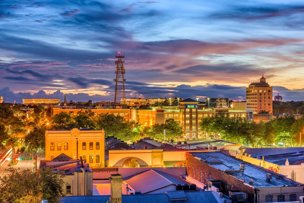 A cityscape at sunset featuring historic and modern buildings, custom homes Gainesville, trees lining the streets, a tall communication tower, and a colorful sky with clouds and fading sunlight.