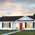 Single-story white house with black roof, black shutters, and a bright red front door. This charming example of custom homes Gainesville features a manicured lawn, neat shrubs, and a pathway leading to the entrance beneath a partly cloudy sunset sky.