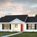A single-story white house with a dark roof, black shutters, and a bright red front door, surrounded by a green lawn and small shrubs—an inviting example of custom homes Gainesville style under a cloudy evening sky.