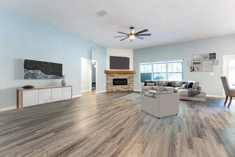 Spacious living room with light blue walls, wood flooring, modern ceiling fan, stone fireplace, large windows, gray sectional sofa, armchair, wall art, and a dining area—perfect for custom homes in North Central Florida.