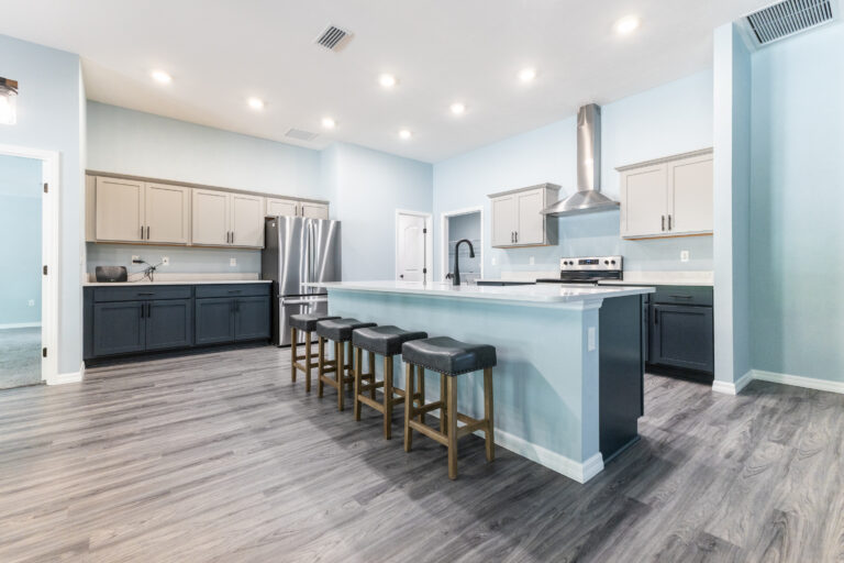 Modern kitchen in custom homes Gainesville with light blue walls, gray cabinets, stainless steel appliances, a large island with four cushioned bar stools, and wood-look flooring under bright recessed lighting.
