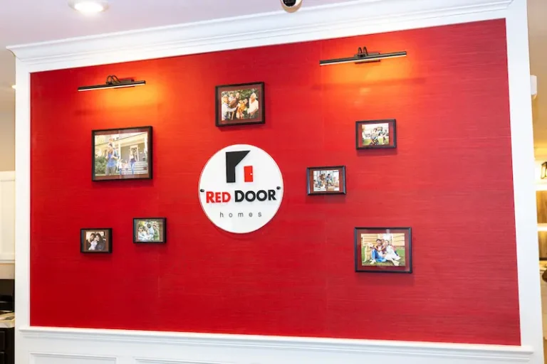 A red wall featuring a white circular Red Door Homes logo in the center, surrounded by framed photos of families, all under two picture lights in a white-trimmed display area.