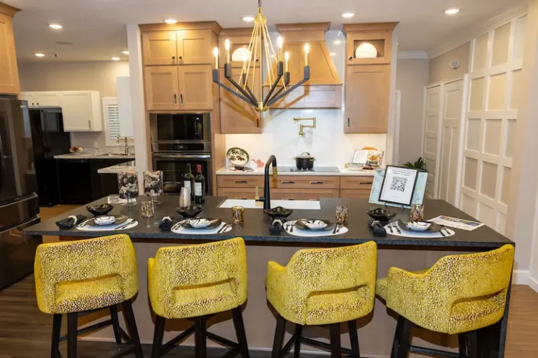 A modern kitchen with wooden cabinets, a black countertop island, four bright yellow barstools, a geometric chandelier, and place settings with wine glasses and plates. The kitchen is well-lit and stylishly decorated.