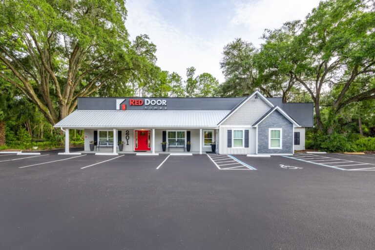A single-story building with a gray exterior, red front door, and Red Door Homes sign. The building is surrounded by trees and has a freshly paved, mostly empty parking lot with marked spaces.