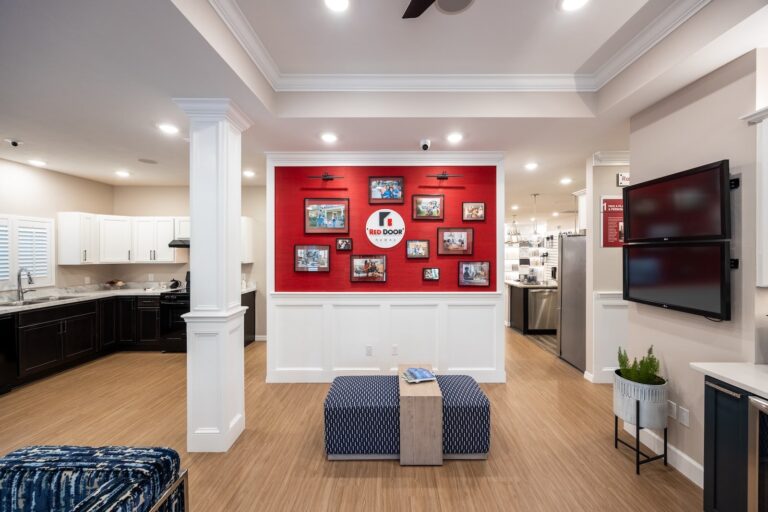 Modern open-concept kitchen and lounge area with wood flooring, white and dark cabinets, a red accent wall displaying framed photos, two ottomans, and a flat-screen TV mounted on the right wall.