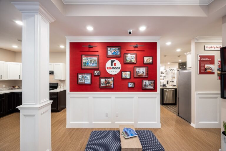 A bright room features a red accent wall displaying multiple framed photos around a Red Door logo. The space has white wainscoting, wood flooring, kitchen area on the left, and a small table with magazines in the foreground.
