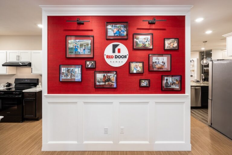 A red accent wall with a “Red Door Homes” sign is decorated with framed family photos, flanked by white paneling below. The wall divides a modern kitchen and living area with wood flooring.