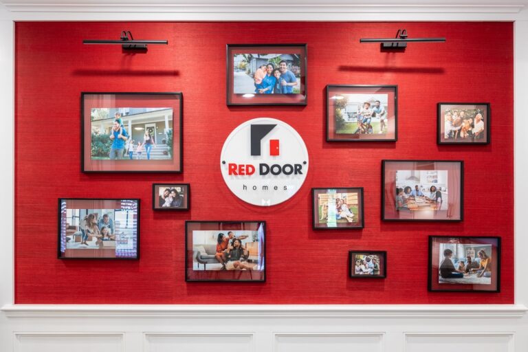 A red wall with the Red Door Homes logo in the center, surrounded by various framed photos of diverse families smiling and spending time together in different home settings.
