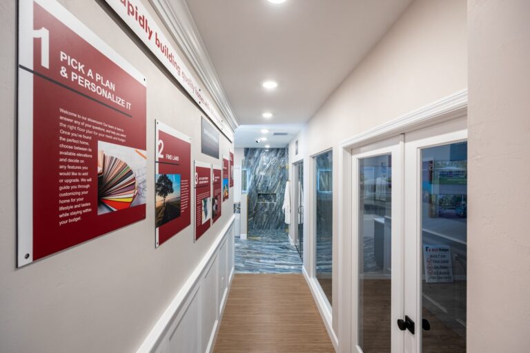 A hallway with informational displays on the left wall, featuring red signs and images about design plans, and glass doors on the right. The floor is wood, and there is bright overhead lighting.