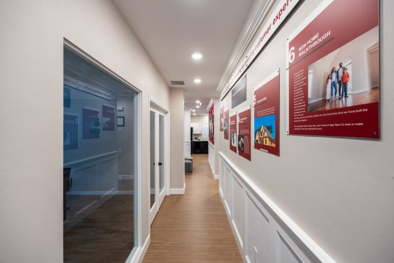 A well-lit hallway with wood flooring, white walls, and framed red informational displays on the right wall. There are glass office windows on the left and a workspace visible at the end of the hall.