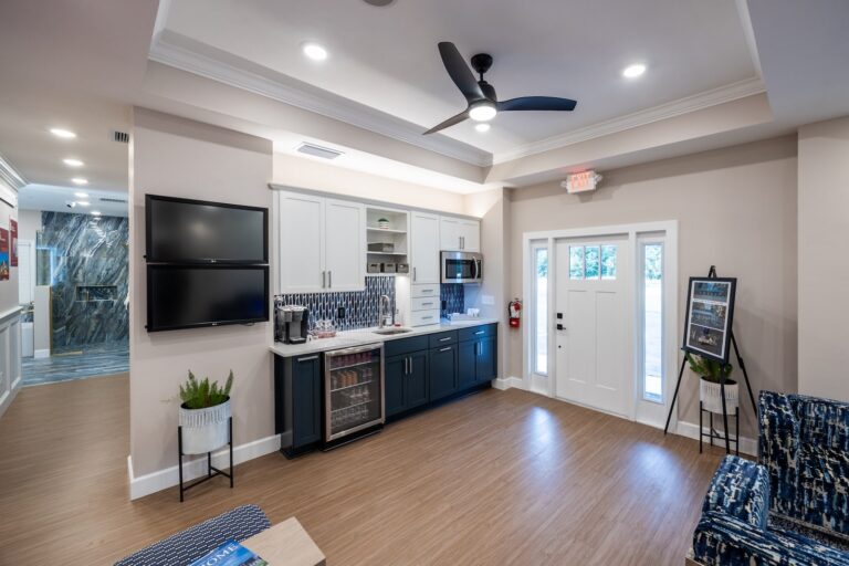 A modern lounge area with a kitchenette, blue and white cabinets, two wall-mounted TVs, a ceiling fan, potted plants, and seating. Large windows and a glass door let in natural light.