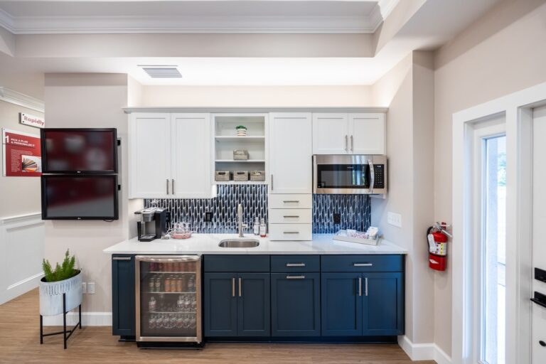 Modern kitchen with white upper cabinets and dark blue lower cabinets, stainless steel sink, microwave, mini-fridge, coffee maker, and tiled backsplash. A fire extinguisher is mounted on the wall near a glass door.
