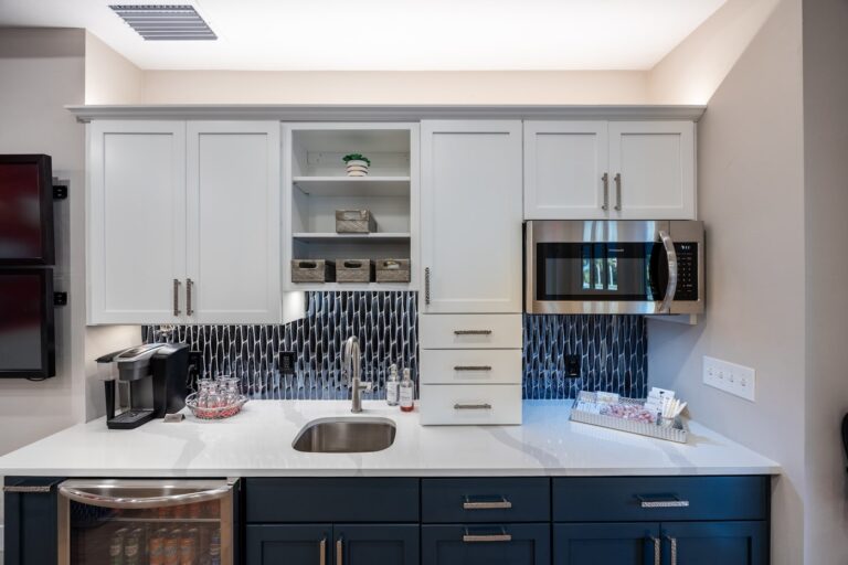 Modern kitchenette with white upper cabinets, a microwave, a small sink, a coffee maker, open shelves with decor, glass jars, and a dark blue lower cabinet. The backsplash features a dark geometric tile pattern.