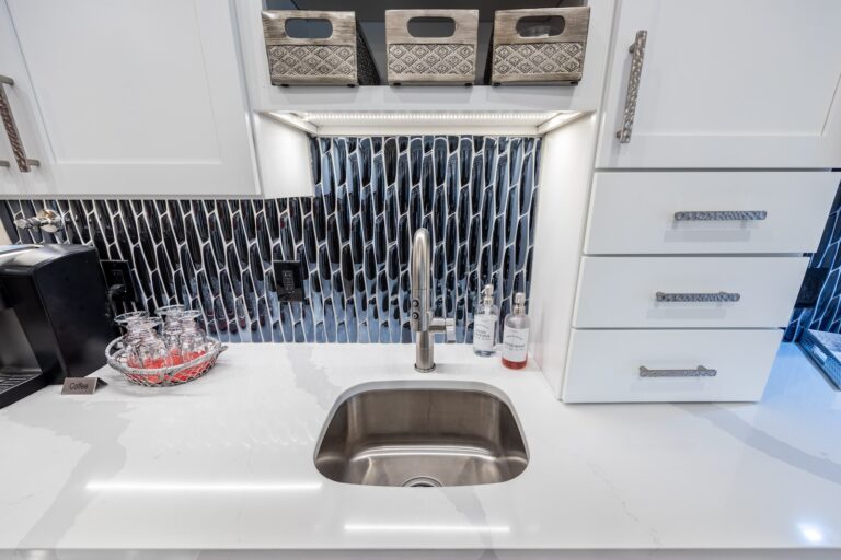 A modern kitchen sink with a silver faucet, white countertop, and black tile backsplash. Three baskets sit in an upper shelf, and soap and glasses are arranged neatly on the counter.