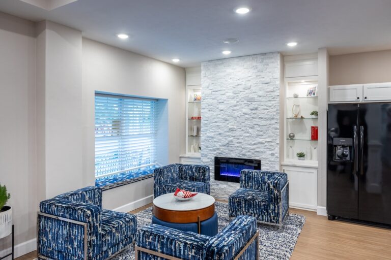 Modern living room with four blue-patterned chairs around a round coffee table, a stone accent wall with electric fireplace, built-in shelves, window with blinds, and a black refrigerator on the right.