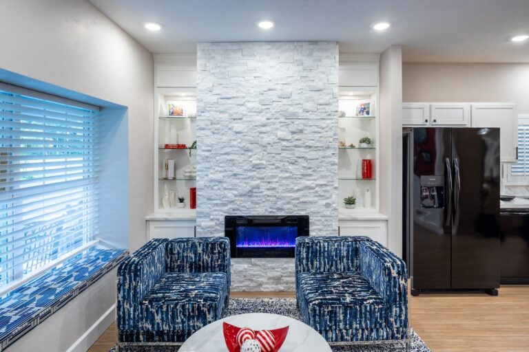 Modern living room with two blue patterned chairs, a round marble table, and a white stone fireplace with an electric fire. Built-in shelves and a black refrigerator are also visible, with natural light from a window on the left.