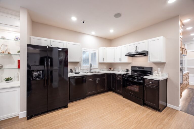 Modern kitchen with black appliances, white upper cabinets, dark lower cabinets, light wood floor, marble countertops, and recessed lighting. A window with white shutters is above the counter.
