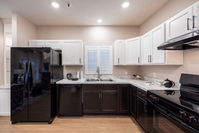 Modern kitchen with black appliances, dark lower cabinets, white upper cabinets, marble countertops, a double sink under a window with shutters, and light wood flooring, all brightly lit by ceiling lights.