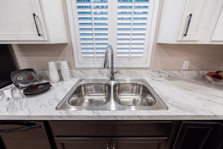 A modern kitchen countertop with a double stainless steel sink, marble surface, white cabinets, and a window with white shutters above the sink. Decor items sit on the countertop.
