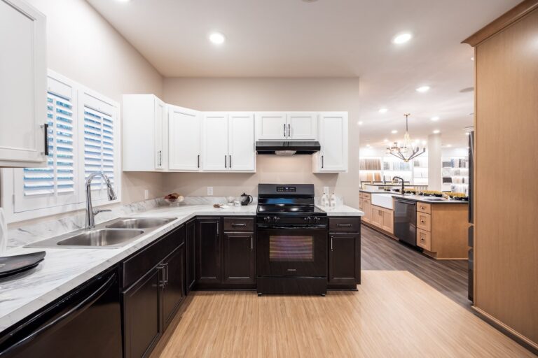 Modern kitchen with white upper cabinets, dark lower cabinets, marble countertops, a black stove and range hood, double sink, and wood flooring. An open dining and living area is visible in the background.
