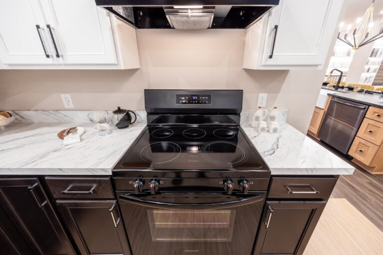 A modern kitchen with a black electric stove and oven set in marble countertops, surrounded by dark lower cabinets and white upper cabinets under bright lighting.