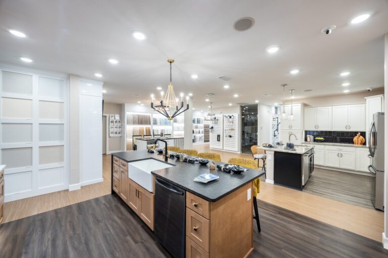 Modern, spacious kitchen with a large island, farmhouse sink, black countertops, and gold-accented chairs; open layout with bright lighting, wood flooring, and white cabinetry throughout.