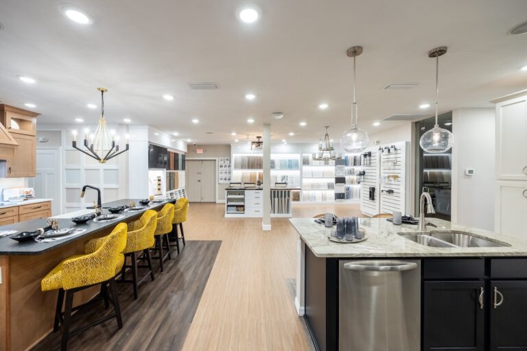 A modern showroom features a kitchen display with yellow bar stools, a large island with a sink, pendant lights, and sample cabinetry and countertop materials showcased on walls. The space is brightly lit and organized.