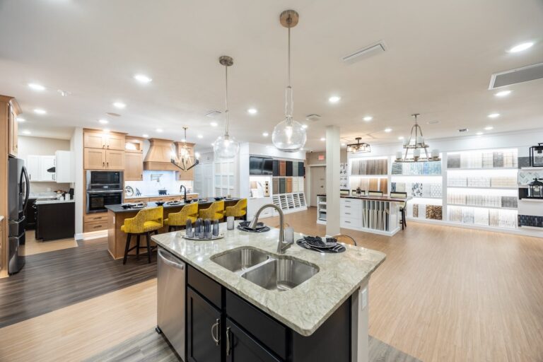 Spacious, modern kitchen showroom with an island sink, pendant lights, yellow chairs at a dining table, wood cabinetry, and wall displays of tiles and materials in a well-lit open layout.