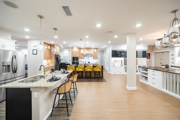 Modern spacious kitchen with an island, yellow bar stools, stainless steel appliances, pendant and recessed lighting, white cabinets, and a dining area with a wood floor and marble countertops.