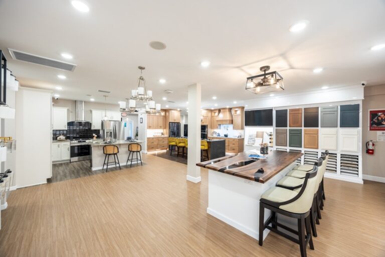 A spacious, modern showroom with a kitchen display, featuring various cabinet styles on the wall, wooden flooring, island counters with bar stools, and bright overhead lighting.
