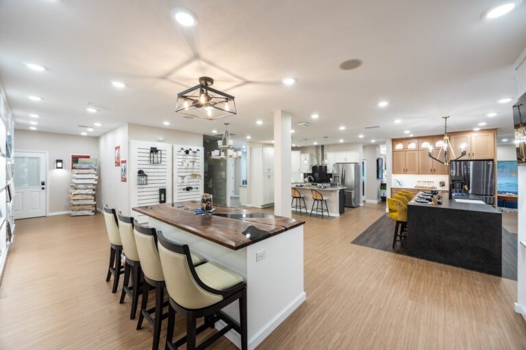 A spacious, modern showroom with a wooden floor, display walls, a counter with stools, and an open kitchen area featuring contemporary lighting and appliances.