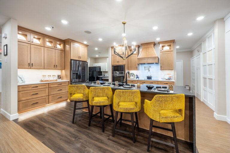 Modern kitchen with wooden cabinets, black appliances, an island with a dark countertop, and five yellow patterned bar stools. Warm lighting and wood flooring create a cozy, inviting atmosphere.