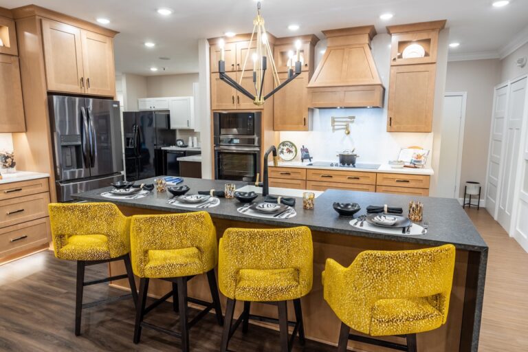 A modern kitchen with wood cabinets, stainless steel appliances, and a dark island countertop. Four yellow upholstered barstools are lined up at the island, which is set with plates and cutlery. A black chandelier hangs above.