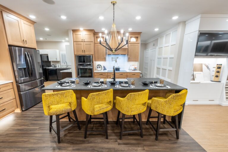 A modern kitchen with wood cabinets, stainless steel appliances, a large island, and four bright yellow textured barstools. The island is set with plates, glasses, and napkins under a stylish chandelier.