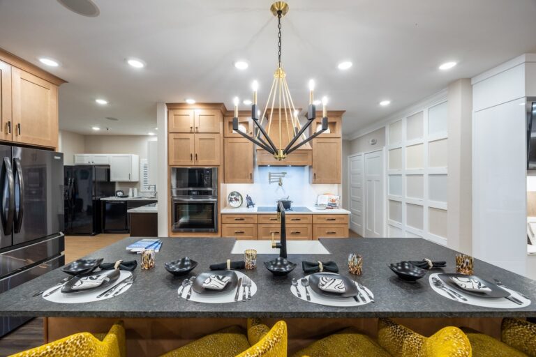 Modern kitchen with light wood cabinets, black appliances, and a large island set for six with black and white plates, bowls, and gold-patterned drinking glasses. A geometric chandelier hangs above the island.