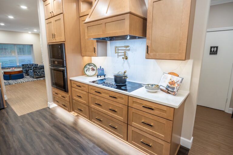 Modern kitchen with wooden cabinets, an electric cooktop, a pot on the stove, cookbook stand, decorative plate, and a double wall oven. White countertops and a light backsplash complete the bright, clean space.