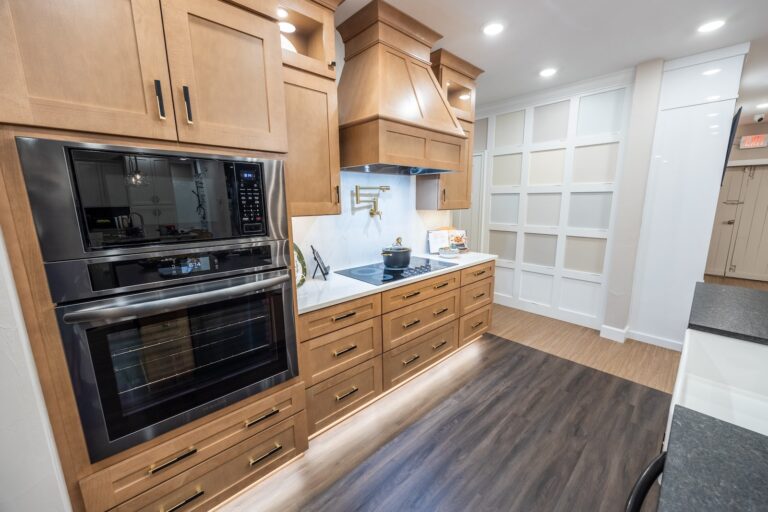 Modern kitchen with wooden cabinets, a built-in double oven, black cooktop, range hood, white backsplash, and under-cabinet lighting. The floor is dark wood, and there’s a white paneled wall in the background.