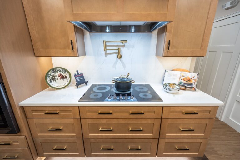 Modern kitchen with light wood cabinets, a black electric cooktop, and a pot on the stove. There is a gold pot filler, a decorative plate, and an open cookbook on the white marble backsplash and countertop.