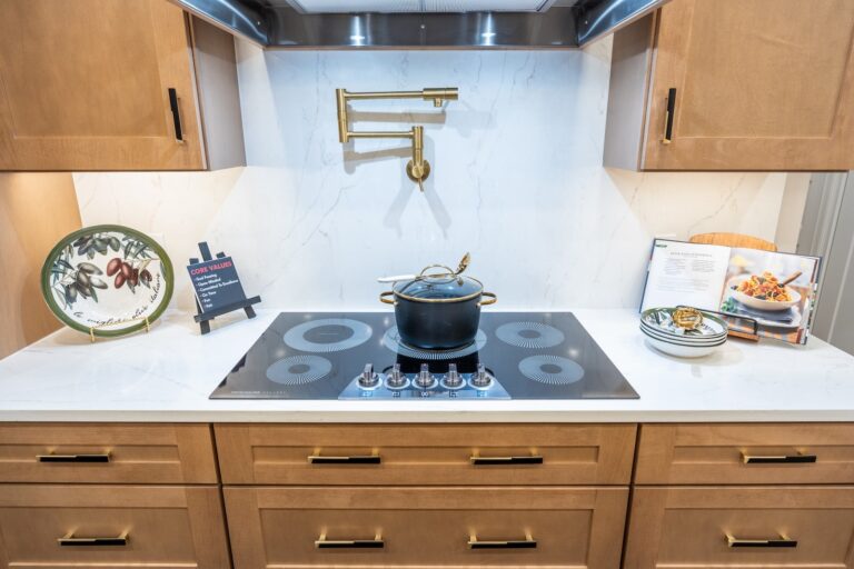 Modern kitchen with a black electric stove, a pot with a ladle on the cooktop, a wall-mounted pot filler, wooden cabinets, a decorative plate, a cookbook on a stand, and a small bowl of food on the counter.