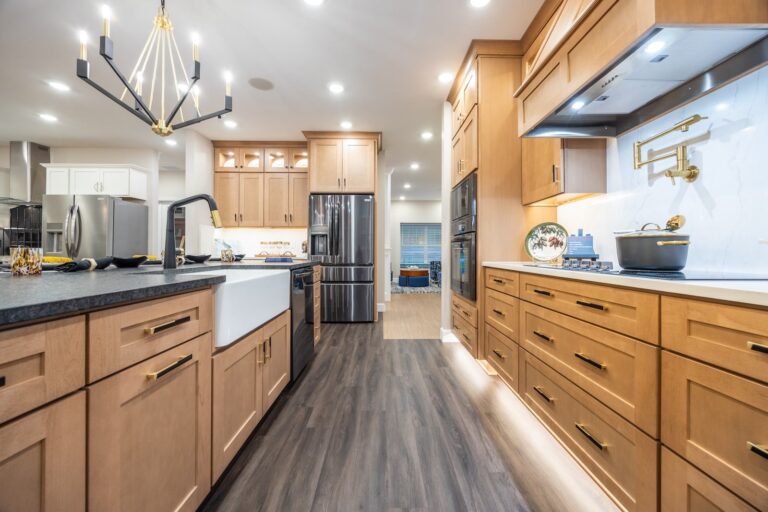 Modern kitchen with light wood cabinets, stainless steel appliances, a farmhouse sink, black countertops, a large chandelier, and under-cabinet lighting. The space is bright and features a wide plank wood floor.
