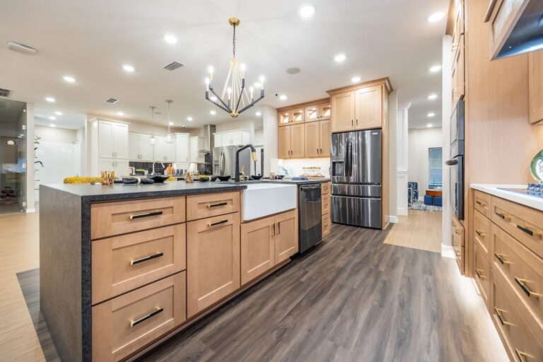 Spacious modern kitchen with wooden cabinets, a large island with a farmhouse sink, black countertops, stainless steel appliances, and a chandelier hanging from the ceiling. The floor is dark wood.