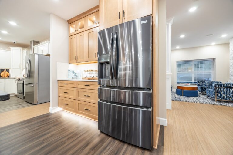 A modern kitchen featuring a stainless steel French door refrigerator, light wood cabinets with under-cabinet lighting, and a wood floor transitioning into an open, well-lit living space.