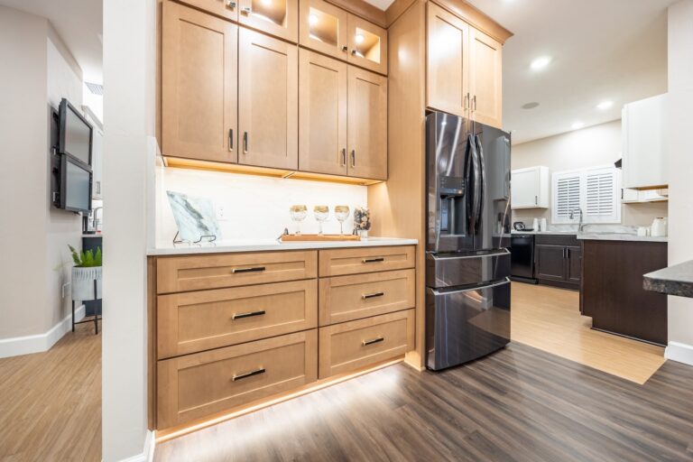 Modern kitchen with light wood cabinets, black refrigerator, under-cabinet lighting, and marble backsplash. The room has a mix of wood and tile flooring, and is clean and well-organized.