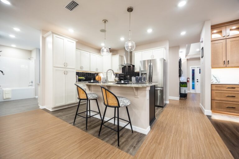 Modern kitchen with white cabinets, a central island with two rattan barstools, stainless steel appliances, pendant lights, and wood-look flooring. A bathroom is visible in the background to the left.