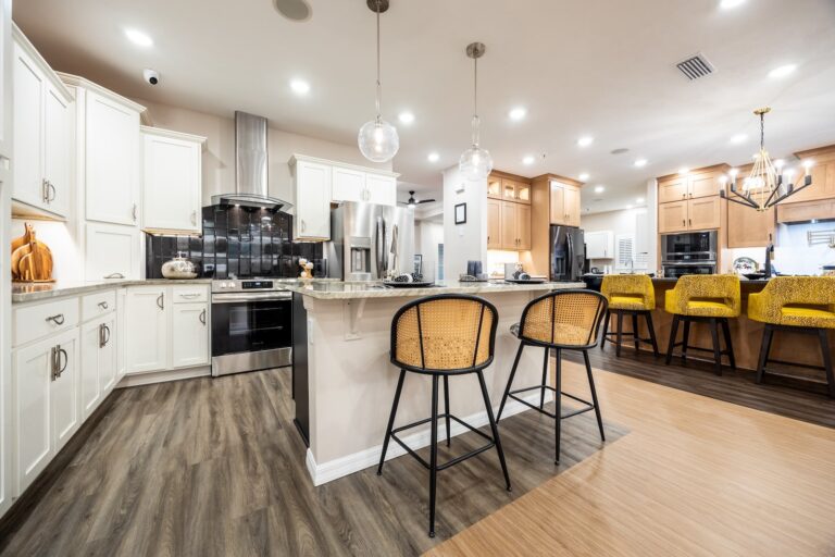 Modern kitchen with white and wood cabinets, stainless steel appliances, a central island with two wicker barstools, pendant lights, and a breakfast bar with four yellow chairs under a contemporary chandelier.