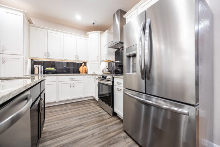 Modern kitchen with white cabinets, stainless steel refrigerator, dishwasher, and oven, black backsplash, marble countertops, and wood flooring. Decorative items and fruit sit on the counters.