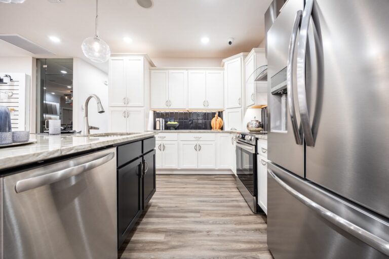 Modern kitchen with white cabinets, stainless steel appliances, a large refrigerator, dishwasher, and oven, black island with sink, and wood-style flooring. Countertops are granite, and decor includes a pumpkin and kitchenware.